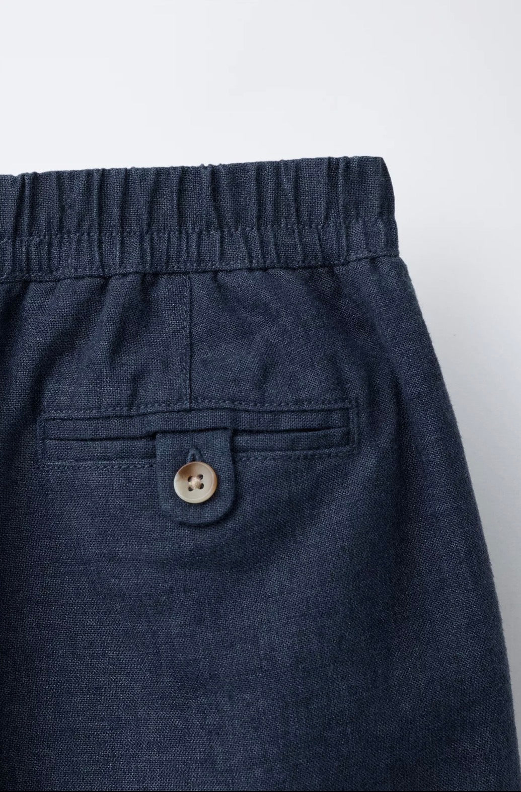 Back detail of navy linen trousers showing elasticated waistband, button-tab back pocket, and woven fabric texture